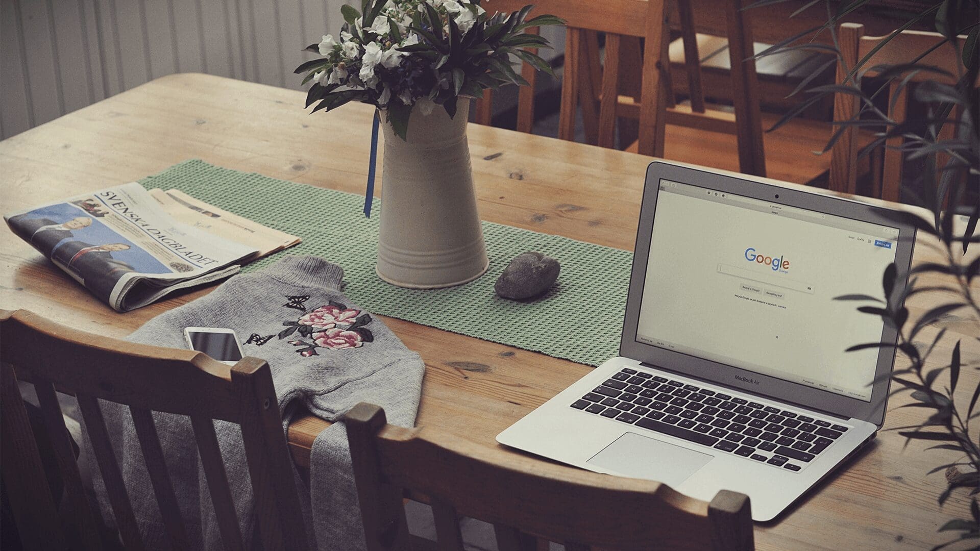 Macbook open to Google search on wooden table with iPhone & newspaper nearby